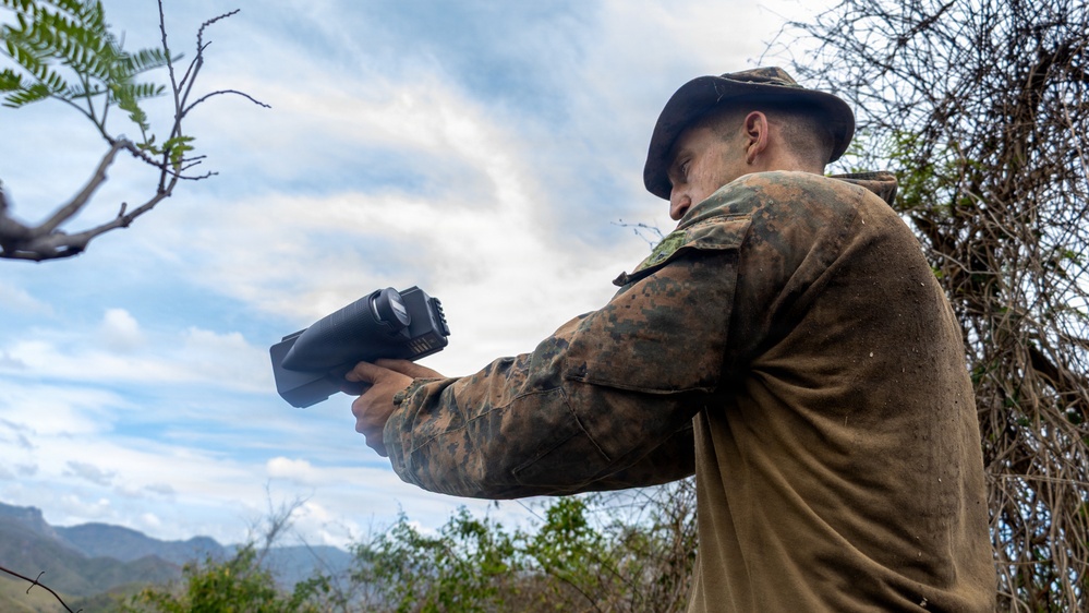 A person wearing military camouflage uniform and a boonie hat is holding and aiming a black handheld optical or thermal imaging device in an outdoor, rugged terrain setting. The individual appears to be conducting reconnaissance or surveillance activity. The background shows mountainous landscape with sparse vegetation and a partly cloudy sky.
