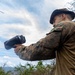 A person wearing military camouflage uniform and a boonie hat is holding and aiming a black handheld optical or thermal imaging device in an outdoor, rugged terrain setting. The individual appears to be conducting reconnaissance or surveillance activity. The background shows mountainous landscape with sparse vegetation and a partly cloudy sky.