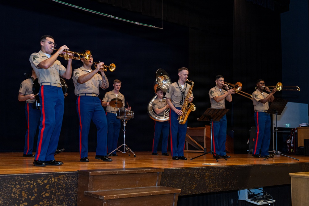 A U.S. Marine Corps band performs on a wooden stage in what appears to be an auditorium or performance hall. The musicians are dressed in standard Marine Corps service uniforms featuring tan shirts, blue trousers with red stripes, and black dress shoes. The ensemble includes brass and percussion instruments in a small concert band formation.
