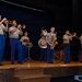 A U.S. Marine Corps band performs on a wooden stage in what appears to be an auditorium or performance hall. The musicians are dressed in standard Marine Corps service uniforms featuring tan shirts, blue trousers with red stripes, and black dress shoes. The ensemble includes brass and percussion instruments in a small concert band formation.