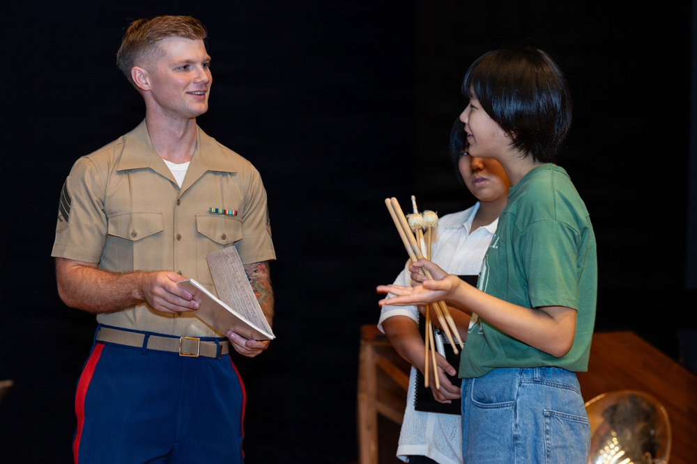 A U.S. Marine in service uniform is engaged in conversation with a young person holding a bundle of drumsticks and mallets, with a second individual visible in the background. The setting appears to be a stage or performance venue with a dark backdrop. The interaction suggests a music outreach or educational engagement event.
