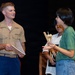 A U.S. Marine in service uniform is engaged in conversation with a young person holding a bundle of drumsticks and mallets, with a second individual visible in the background. The setting appears to be a stage or performance venue with a dark backdrop. The interaction suggests a music outreach or educational engagement event.