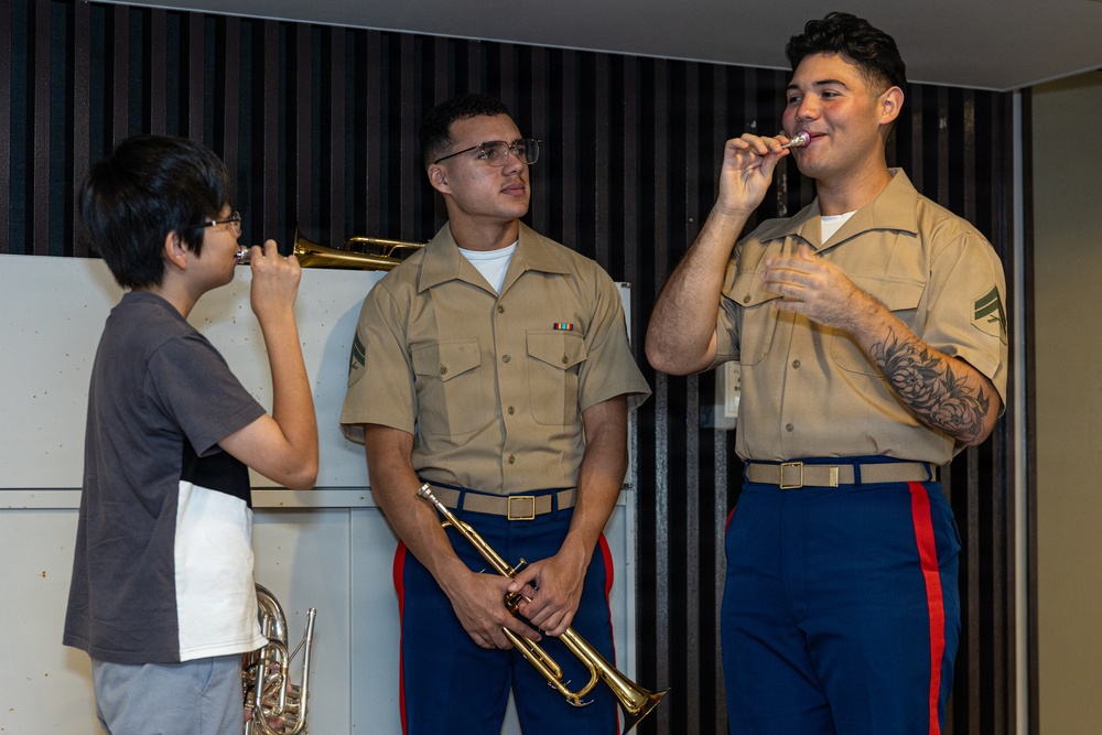Two U.S. Marines in service uniforms interact with a young civilian student in what appears to be a music room or rehearsal space. The Marines are demonstrating trumpet mouthpiece technique to the child, who is attempting to buzz into a mouthpiece. This appears to be an outreach or educational event where military musicians are engaging with youth.