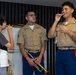 Two U.S. Marines in service uniforms interact with a young civilian student in what appears to be a music room or rehearsal space. The Marines are demonstrating trumpet mouthpiece technique to the child, who is attempting to buzz into a mouthpiece. This appears to be an outreach or educational event where military musicians are engaging with youth.