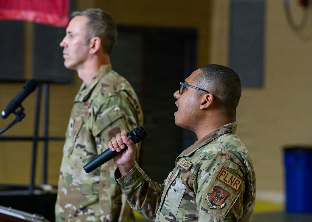 Two military personnel in OCP (Operational Camouflage Pattern) uniforms are participating in what appears to be a formal military ceremony or briefing indoors. The foreground individual is speaking into a handheld microphone while the background individual stands at attention near a podium with a microphone stand. The setting appears to be a gymnasium or large indoor military facility.