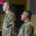 Two military personnel in OCP (Operational Camouflage Pattern) uniforms are participating in what appears to be a formal military ceremony or briefing indoors. The foreground individual is speaking into a handheld microphone while the background individual stands at attention near a podium with a microphone stand. The setting appears to be a gymnasium or large indoor military facility.