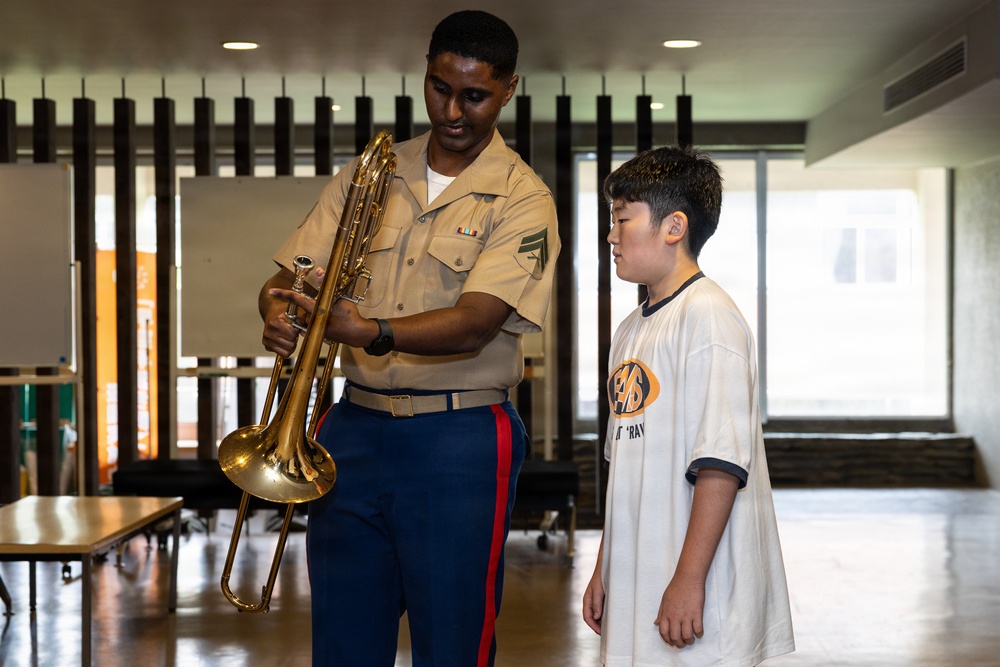 A U.S. Marine in service uniform is showing a trombone to a young boy in a casual white t-shirt, likely during an educational outreach or community engagement event. The Marine appears to be explaining or demonstrating the brass instrument to the attentive youth. The setting appears to be an indoor multipurpose room or community hall.