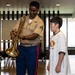 A U.S. Marine in service uniform is showing a trombone to a young boy in a casual white t-shirt, likely during an educational outreach or community engagement event. The Marine appears to be explaining or demonstrating the brass instrument to the attentive youth. The setting appears to be an indoor multipurpose room or community hall.