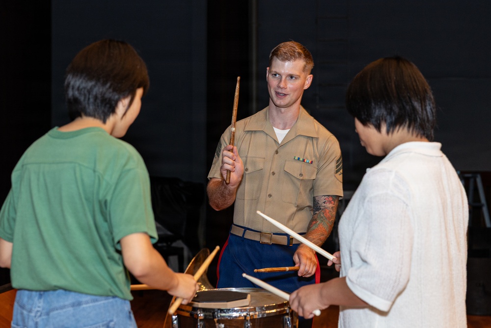 A U.S. Marine in service uniform is teaching two young students how to play drums. The Marine, holding drumsticks, appears to be giving instruction while the two children practice on snare drums. The setting appears to be a stage or performance hall, suggesting a community outreach or music education event.