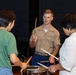 A U.S. Marine in service uniform is teaching two young students how to play drums. The Marine, holding drumsticks, appears to be giving instruction while the two children practice on snare drums. The setting appears to be a stage or performance hall, suggesting a community outreach or music education event.