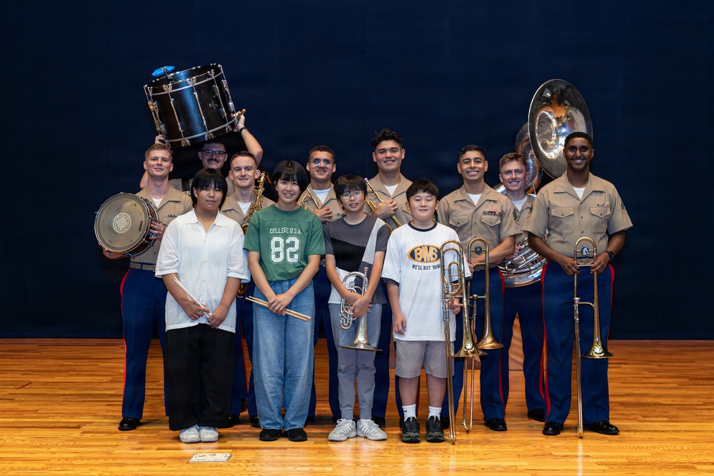 A group photo on a wooden stage featuring several U.S. Marines in tan/khaki service uniforms alongside four younger civilians, likely students. The group is posing with various musical instruments including drums, a sousaphone/tuba, a trombone, a trumpet, and a saxophone. This appears to be a music outreach or educational event involving the Marine Corps band.