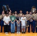 A group photo on a wooden stage featuring several U.S. Marines in tan/khaki service uniforms alongside four younger civilians, likely students. The group is posing with various musical instruments including drums, a sousaphone/tuba, a trombone, a trumpet, and a saxophone. This appears to be a music outreach or educational event involving the Marine Corps band.