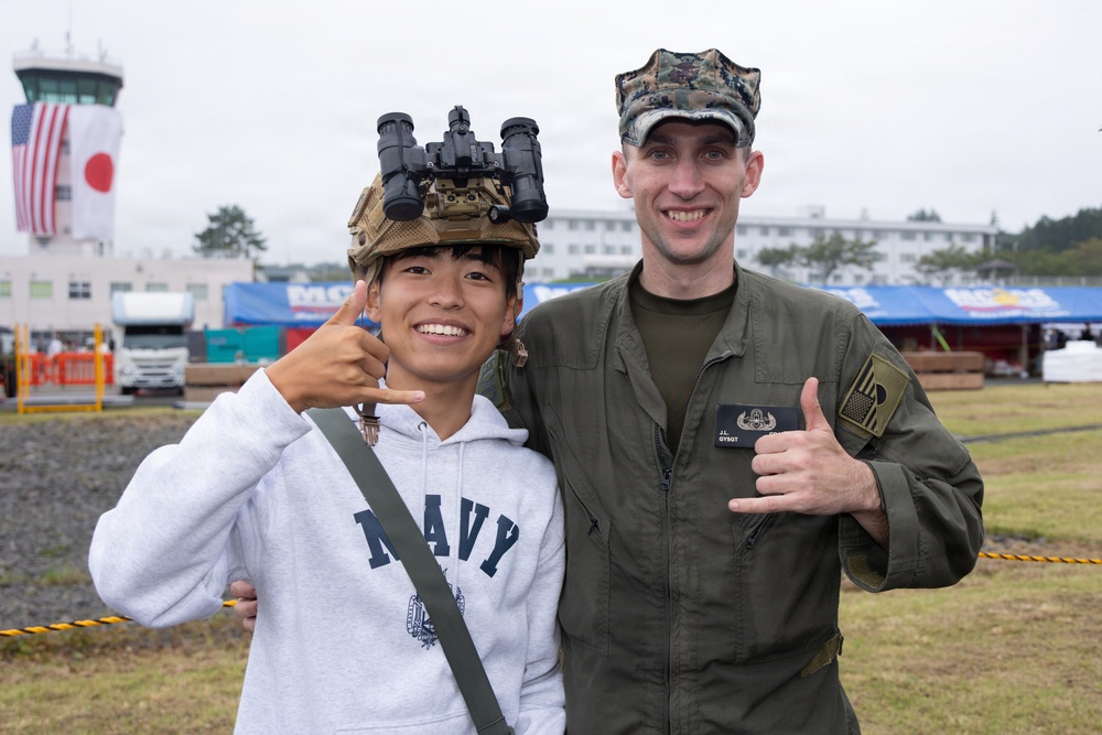 Two individuals pose together at what appears to be a military air show or open base event, likely in Japan based on the visible Japanese and American flags in the background. A young civilian wearing a Navy sweatshirt has a military tactical helmet with night-vision goggles placed on his head, while a U.S. Marine in a flight suit and cover (cap) gives a thumbs up. Both are smiling and making hand gestures in a friendly, casual photo opportunity.