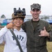 Two individuals pose together at what appears to be a military air show or open base event, likely in Japan based on the visible Japanese and American flags in the background. A young civilian wearing a Navy sweatshirt has a military tactical helmet with night-vision goggles placed on his head, while a U.S. Marine in a flight suit and cover (cap) gives a thumbs up. Both are smiling and making hand gestures in a friendly, casual photo opportunity.
