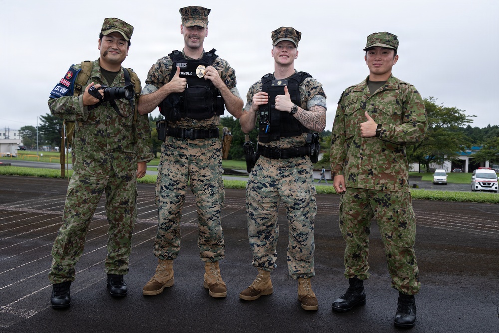 Four military personnel pose together on a wet pavement area, appearing to be at a joint military installation. Two individuals wear U.S. Marine Corps MARPAT digital camouflage with black tactical vests, while two others wear Japanese Ground Self-Defense Force (JGSDF) camouflage uniforms. The group appears to be participating in a joint military exercise or cooperative event.