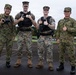 Four military personnel pose together on a wet pavement area, appearing to be at a joint military installation. Two individuals wear U.S. Marine Corps MARPAT digital camouflage with black tactical vests, while two others wear Japanese Ground Self-Defense Force (JGSDF) camouflage uniforms. The group appears to be participating in a joint military exercise or cooperative event.