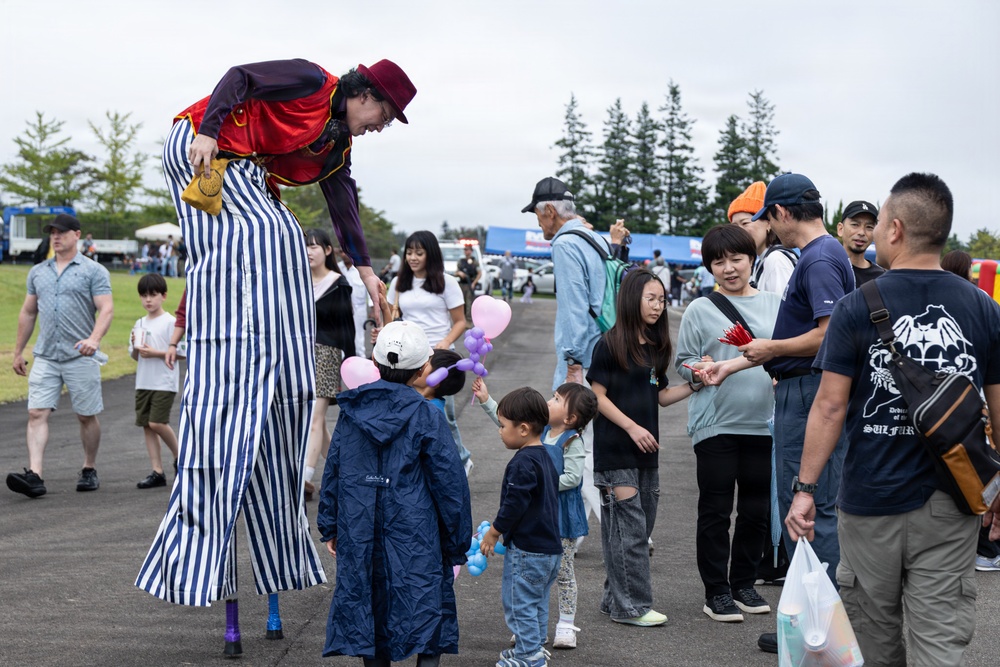 A stilt-walker performer dressed in a colorful circus costume with blue and white striped pants and a red cape is interacting with children and families at an outdoor festival. The performer is bending down to engage with young children, one of whom is holding balloon animals. The event appears to be a community or air base festival held on a paved outdoor area with trees and vendor tents visible in the background.