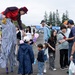 A stilt-walker performer dressed in a colorful circus costume with blue and white striped pants and a red cape is interacting with children and families at an outdoor festival. The performer is bending down to engage with young children, one of whom is holding balloon animals. The event appears to be a community or air base festival held on a paved outdoor area with trees and vendor tents visible in the background.