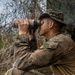 A military service member in woodland camouflage uniform and boonie hat is using binoculars to observe a distant area from a concealed position. The individual is positioned near natural brush and a camouflage netting structure, suggesting a surveillance or reconnaissance operation. The setting appears to be a field training exercise or operational deployment in a vegetated outdoor environment.