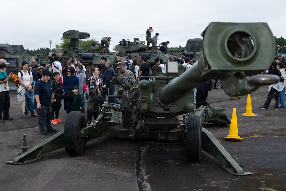 A military open day or public exhibition event where civilians, including families with children, are viewing military hardware up close. A large green artillery piece (likely an M198 or M777 howitzer) dominates the foreground, with armored vehicles and tanks visible in the background. At least one uniformed military personnel is present, appearing to explain or demonstrate equipment to visitors.