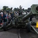 A military open day or public exhibition event where civilians, including families with children, are viewing military hardware up close. A large green artillery piece (likely an M198 or M777 howitzer) dominates the foreground, with armored vehicles and tanks visible in the background. At least one uniformed military personnel is present, appearing to explain or demonstrate equipment to visitors.