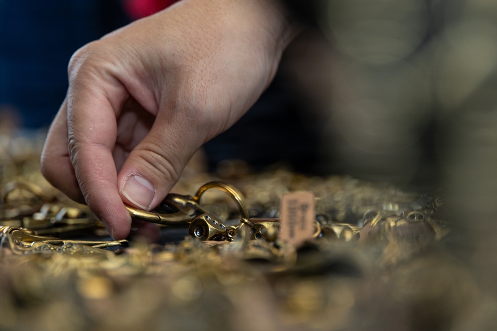 A hand is reaching into a large pile of gold-toned jewelry items, appearing to pick up or examine a specific piece. The collection includes rings, chains, clasps, and other jewelry components spread across a surface. At least one item has a small price or identification tag attached, suggesting a retail, auction, or resale context.