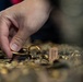 A hand is reaching into a large pile of gold-toned jewelry items, appearing to pick up or examine a specific piece. The collection includes rings, chains, clasps, and other jewelry components spread across a surface. At least one item has a small price or identification tag attached, suggesting a retail, auction, or resale context.