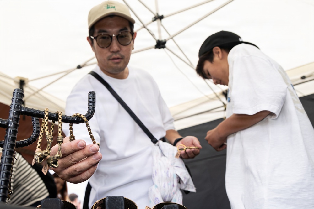 Two individuals are browsing jewelry or accessories at an outdoor market stall under a white canopy tent. The foreground person, a man wearing a beige cap, round glasses, and a white t-shirt with a crossbody bag, is examining gold-toned chain jewelry displayed on a black rack. A second person in a white t-shirt and black cap is bent over, inspecting a small item in the background.