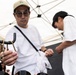 Two individuals are browsing jewelry or accessories at an outdoor market stall under a white canopy tent. The foreground person, a man wearing a beige cap, round glasses, and a white t-shirt with a crossbody bag, is examining gold-toned chain jewelry displayed on a black rack. A second person in a white t-shirt and black cap is bent over, inspecting a small item in the background.