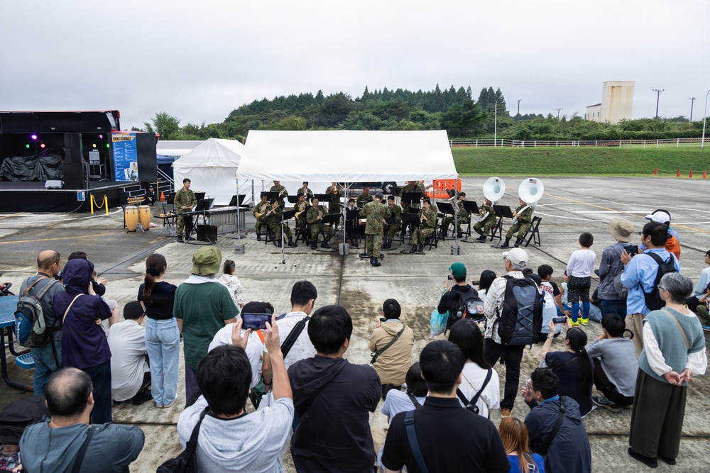 A military band performs outdoors on a paved area, likely a base or airfield, before a crowd of civilian spectators. The musicians are dressed in military uniforms and are playing brass and percussion instruments. The event appears to be a public open-day or community outreach performance held under overcast skies.