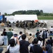 A military band performs outdoors on a paved area, likely a base or airfield, before a crowd of civilian spectators. The musicians are dressed in military uniforms and are playing brass and percussion instruments. The event appears to be a public open-day or community outreach performance held under overcast skies.
