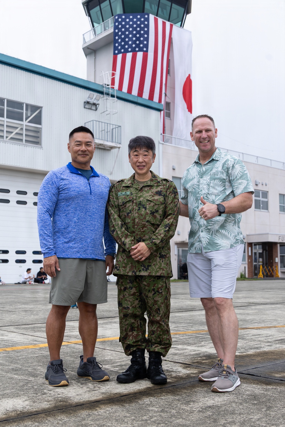 Three men pose for a photo on what appears to be a military airfield tarmac. The central figure is wearing Japanese Self-Defense Force camouflage uniform, flanked by two men in civilian casual attire. Behind them is an air traffic control tower displaying both the United States flag and the Japanese flag side by side.
