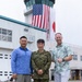 Three men pose for a photo on what appears to be a military airfield tarmac. The central figure is wearing Japanese Self-Defense Force camouflage uniform, flanked by two men in civilian casual attire. Behind them is an air traffic control tower displaying both the United States flag and the Japanese flag side by side.