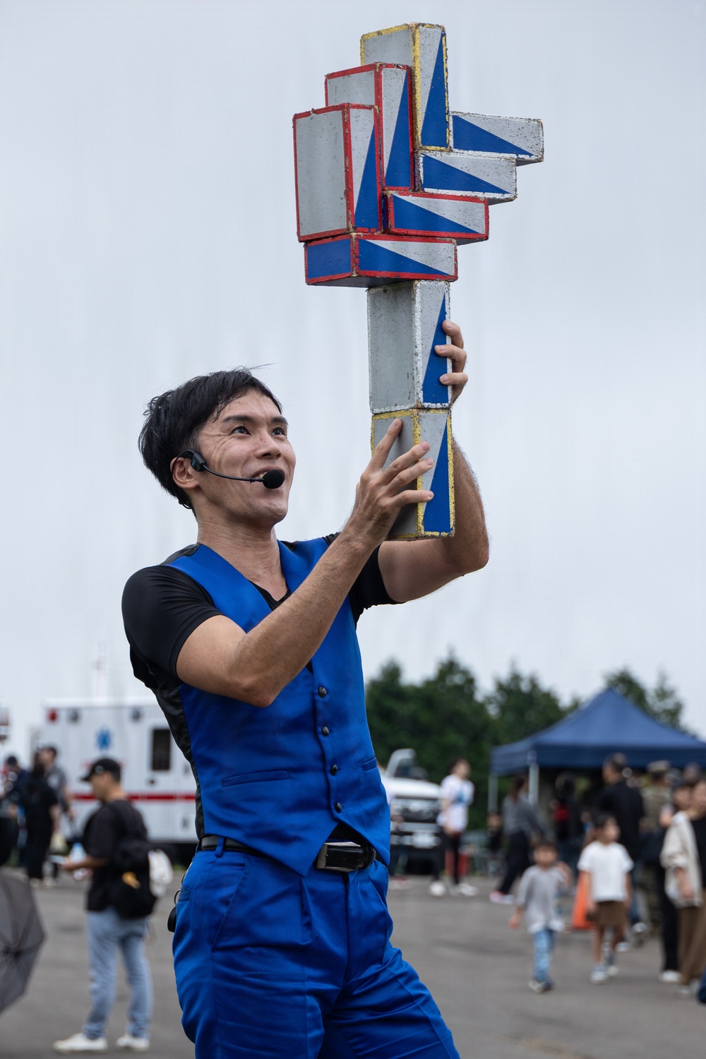 A male performer dressed in a blue vest and blue pants holds up a large geometric prop made of interlocking rectangular blocks painted in blue, white, red, and yellow. He is wearing a headset microphone and appears to be mid-performance at an outdoor public event. The background shows a crowd of spectators, trees, a blue canopy tent, and what appears to be an ambulance.