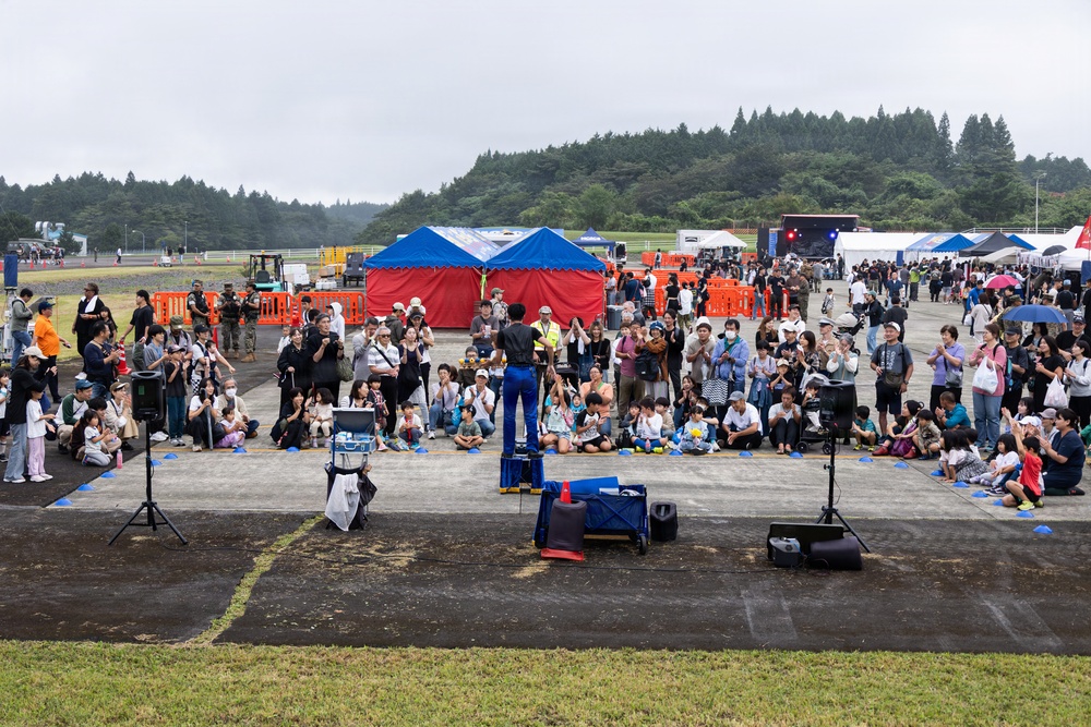 An outdoor public event or festival is taking place on a large paved grounds, likely in Japan, on an overcast or rainy day. A performer stands with their back to the camera, facing a large seated and standing crowd of attendees including many children and families. The venue features multiple tents, vendor stalls, a stage with lighting in the background, and surrounding forested hills.