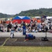 An outdoor public event or festival is taking place on a large paved grounds, likely in Japan, on an overcast or rainy day. A performer stands with their back to the camera, facing a large seated and standing crowd of attendees including many children and families. The venue features multiple tents, vendor stalls, a stage with lighting in the background, and surrounding forested hills.