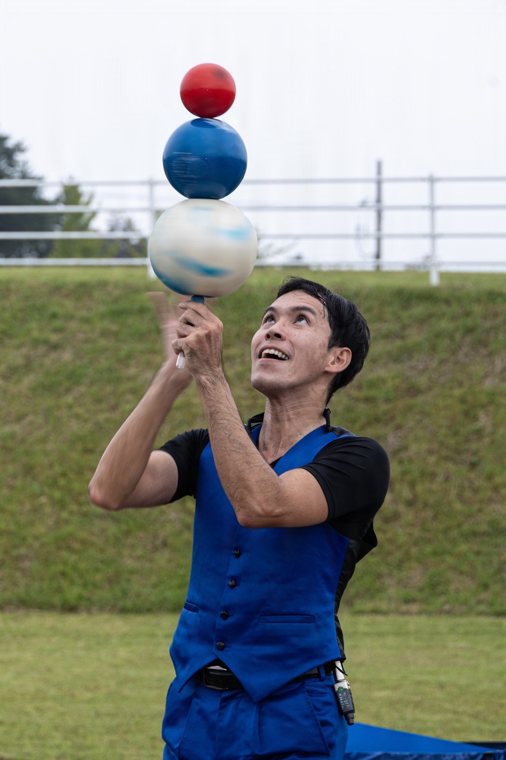 A male performer is balancing three stacked spherical balls on a short stick or handle held in both hands, gazing upward with a smile. He is dressed in a blue vest over a black shirt with blue trousers, performing outdoors on a grassy area. The three balls are stacked in descending size order: a large white/blue marbled ball at the bottom, a medium solid blue ball in the middle, and a small solid red ball on top.