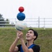 A male performer is balancing three stacked spherical balls on a short stick or handle held in both hands, gazing upward with a smile. He is dressed in a blue vest over a black shirt with blue trousers, performing outdoors on a grassy area. The three balls are stacked in descending size order: a large white/blue marbled ball at the bottom, a medium solid blue ball in the middle, and a small solid red ball on top.