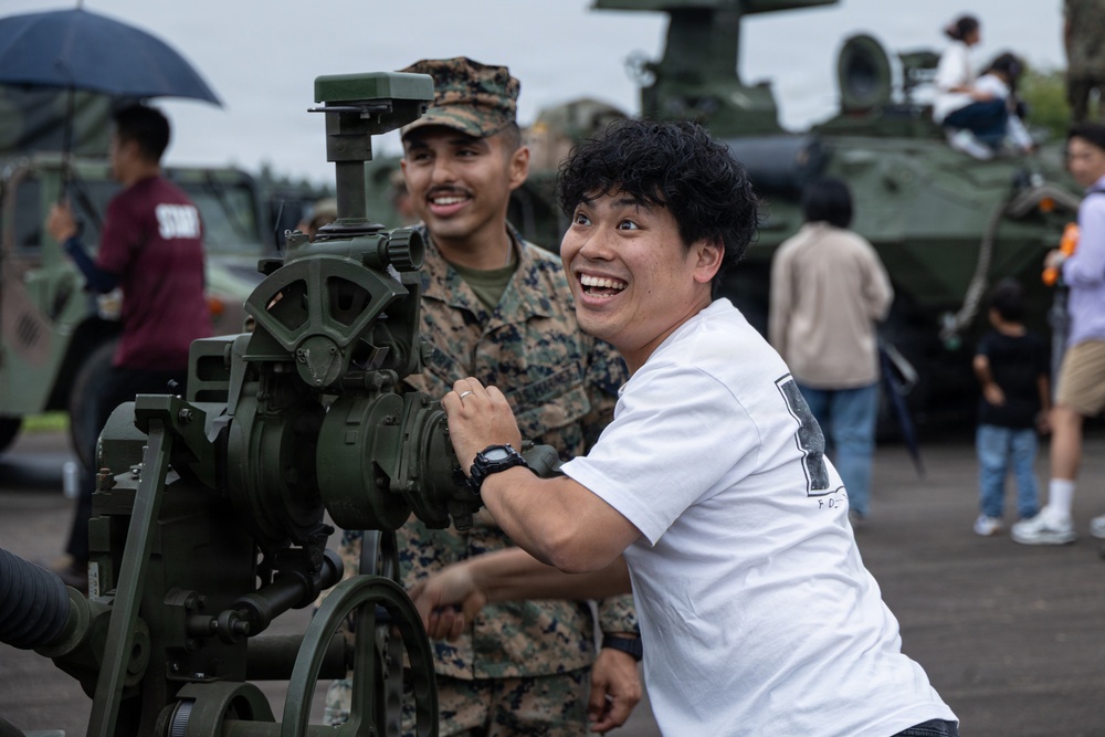 A civilian man in a white t-shirt enthusiastically interacts with a military artillery piece or mortar at what appears to be a public military open day or air show. A U.S. Marine in woodland MARPAT camouflage uniform and cover stands behind him, smiling. The background shows additional military vehicles, including what appears to be a LAV or similar armored vehicle, with other civilians exploring the equipment.