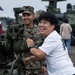 A civilian man in a white t-shirt enthusiastically interacts with a military artillery piece or mortar at what appears to be a public military open day or air show. A U.S. Marine in woodland MARPAT camouflage uniform and cover stands behind him, smiling. The background shows additional military vehicles, including what appears to be a LAV or similar armored vehicle, with other civilians exploring the equipment.