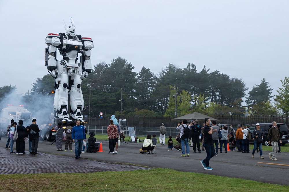A large white mecha/robot statue or prop, resembling a design from the anime series Patlabor or a similar mecha franchise, is displayed at an outdoor public event. Crowds of people, appearing to be in Japan based on context clues, gather around the towering figure to observe and photograph it. Smoke or fog effects surround the base of the robot, adding to the dramatic presentation.