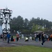 A large white mecha/robot statue or prop, resembling a design from the anime series Patlabor or a similar mecha franchise, is displayed at an outdoor public event. Crowds of people, appearing to be in Japan based on context clues, gather around the towering figure to observe and photograph it. Smoke or fog effects surround the base of the robot, adding to the dramatic presentation.