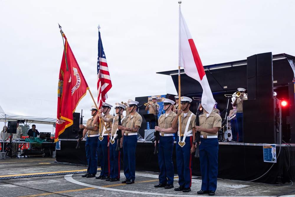 A U.S. Marine Corps color guard stands at attention during a formal ceremony, holding three flags: a red Marine Corps unit flag (partially reading 'Camp Fuji'), the U.S. flag, and the Japanese national flag. The Marines are in dress uniform with tan shirts, blue trousers, white covers, and white rifle slings. A military band performs on a stage in the background, suggesting a joint U.S.-Japan military event.