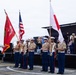 A U.S. Marine Corps color guard stands at attention during a formal ceremony, holding three flags: a red Marine Corps unit flag (partially reading 'Camp Fuji'), the U.S. flag, and the Japanese national flag. The Marines are in dress uniform with tan shirts, blue trousers, white covers, and white rifle slings. A military band performs on a stage in the background, suggesting a joint U.S.-Japan military event.