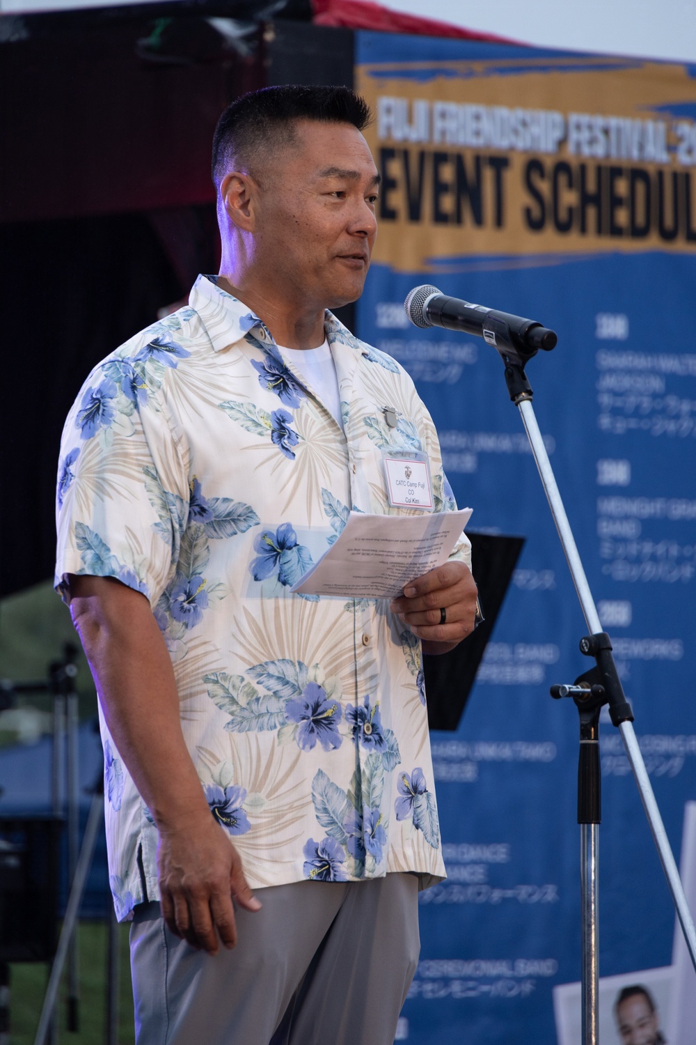 A man in a floral Hawaiian shirt is speaking at a microphone podium while holding papers, at an event called the 'Fuji Friendship Festival.' He is wearing a name badge and appears to be delivering a speech or announcement. The backdrop features a blue event schedule banner with both English and Japanese text.