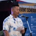 A man in a floral Hawaiian shirt is speaking at a microphone podium while holding papers, at an event called the 'Fuji Friendship Festival.' He is wearing a name badge and appears to be delivering a speech or announcement. The backdrop features a blue event schedule banner with both English and Japanese text.