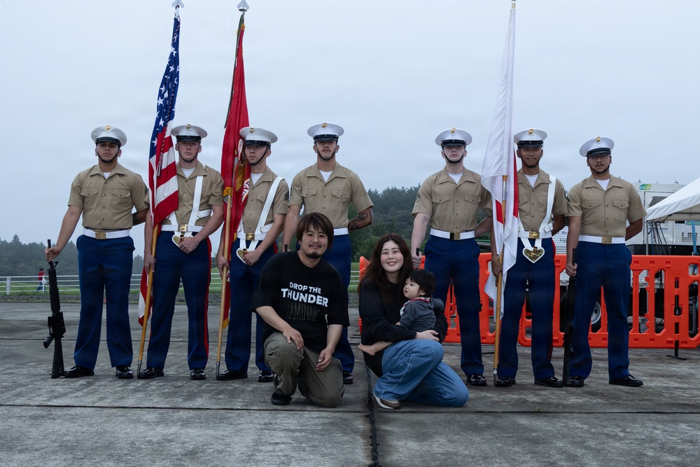 A color guard of seven U.S. Marines in dress uniform stands at attention holding flags, including the American flag, a Marine Corps flag, and what appears to be a Japanese flag. A civilian family of three — a man, a woman holding a young child — poses in front of the color guard for a photograph. The setting appears to be an outdoor military event or air show, likely at a U.S. military installation in Japan.