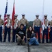 A color guard of seven U.S. Marines in dress uniform stands at attention holding flags, including the American flag, a Marine Corps flag, and what appears to be a Japanese flag. A civilian family of three — a man, a woman holding a young child — poses in front of the color guard for a photograph. The setting appears to be an outdoor military event or air show, likely at a U.S. military installation in Japan.