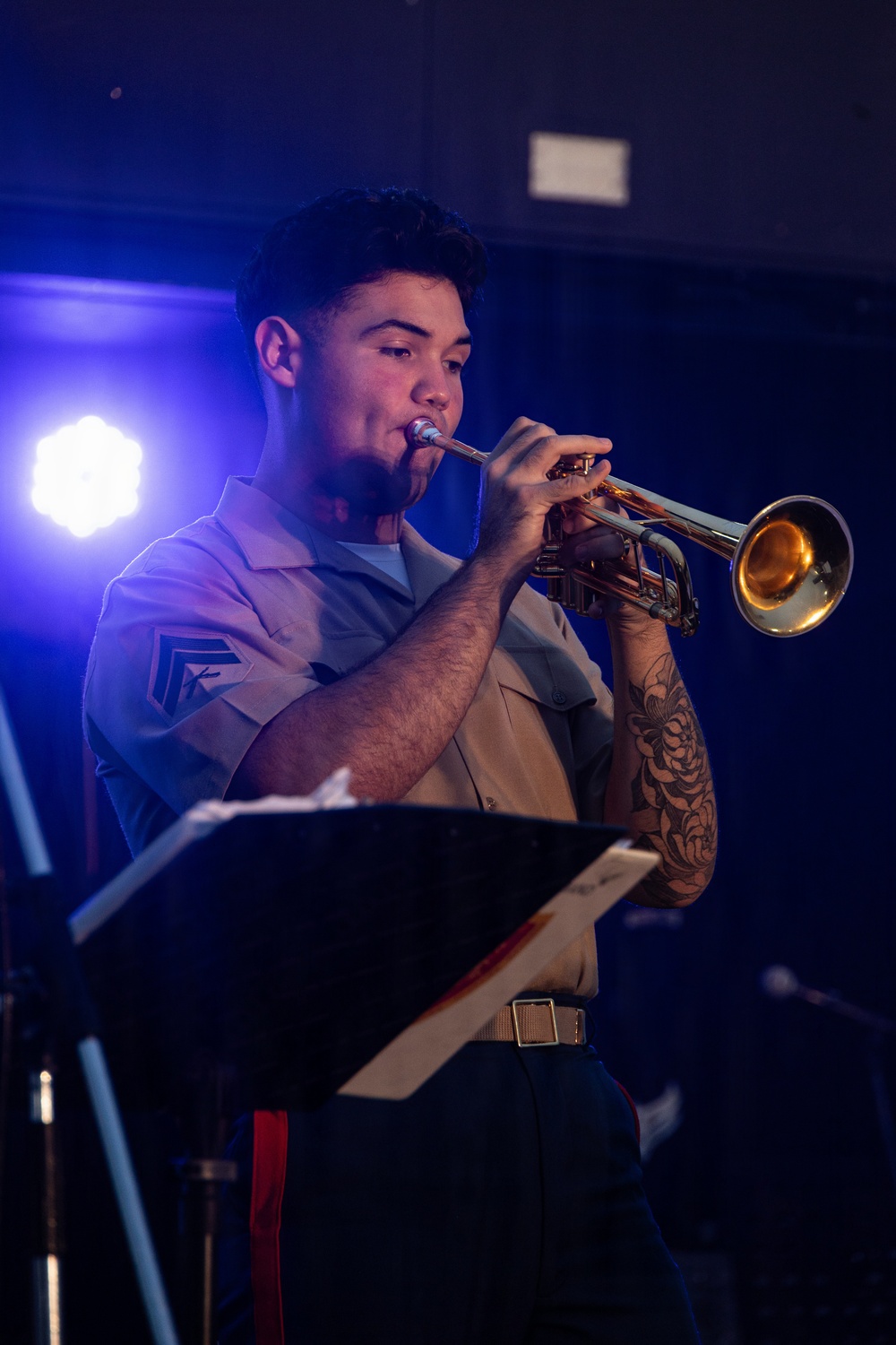 A military service member in a tan/khaki uniform is playing a brass trumpet during what appears to be an indoor performance or concert. The performer stands at a music stand under blue stage lighting, with sheet music visible on the stand. A floral tattoo sleeve is visible on the performer's right arm.