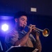 A military service member in a tan/khaki uniform is playing a brass trumpet during what appears to be an indoor performance or concert. The performer stands at a music stand under blue stage lighting, with sheet music visible on the stand. A floral tattoo sleeve is visible on the performer's right arm.